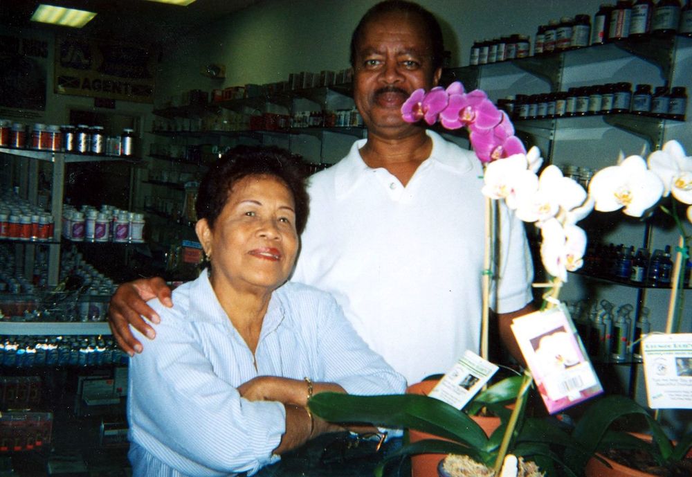 Hyacinth and Merton Cooke, founders of Jamaica Herbal, standing together inside the original Lauderdale Lakes store with orchids and supplement shelves behind them